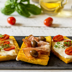 Gegrillte Polenta-Schnitten mit Stracchino, geschmorten Kirschtomaten, Prosciutto und Oliven auf einer Platte