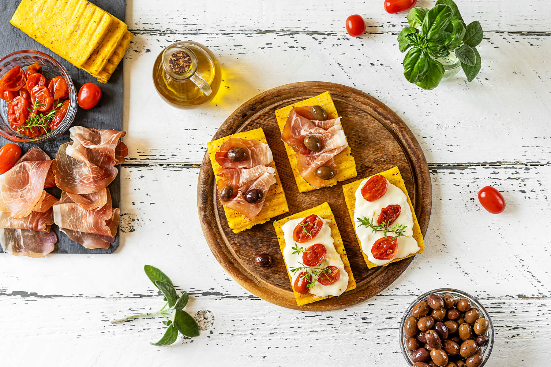 Gegrillte Polenta-Schnitten auf einem Holzbrett, teils mit Prosciutto und Oliven, teils mit Käse und Tomaten belegt