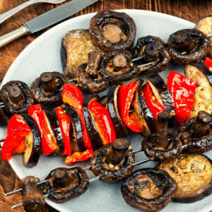 Bunte Gemüsespieße vom Grill mit Champignons, Aubergine und roter Paprika auf hellem Teller, daneben Gabel und Messer auf Holzuntergrund