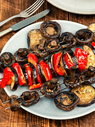 Bunte Gemüsespieße vom Grill mit Champignons, Aubergine und roter Paprika auf hellem Teller, daneben Gabel und Messer auf Holzuntergrund