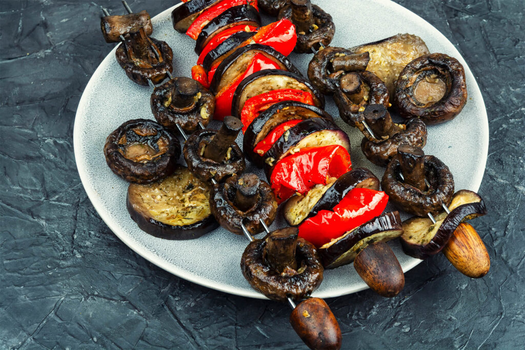 Bunte Gemüsespieße vom Grill mit Champignons, Auberginenscheiben und roter Paprika auf hellem Teller vor dunklem Hintergrund