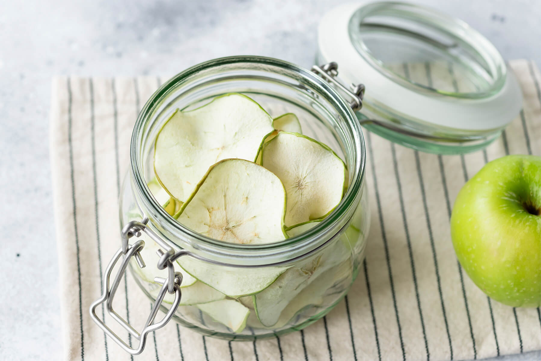 Getrocknete Apfelchips in einem Bügelglas, daneben ein grüner Apfel