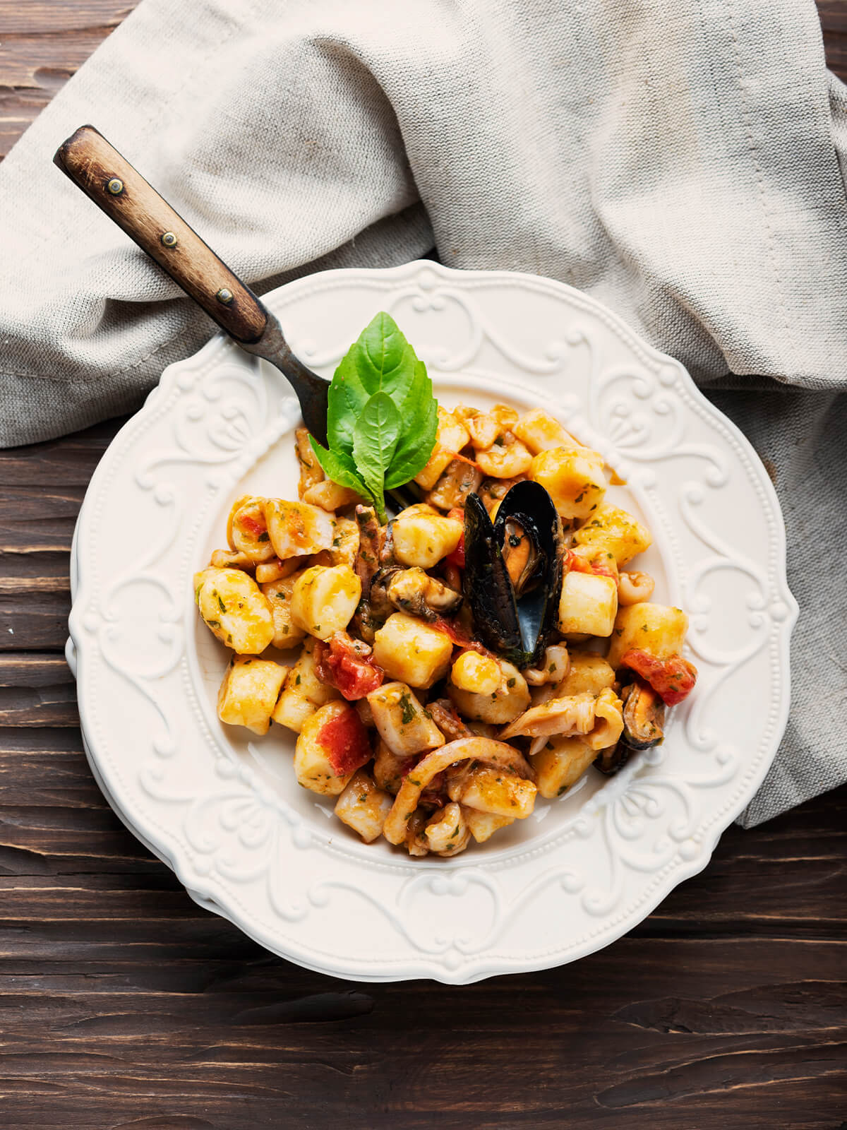 Blick von oben auf einen weißen Teller mit verziertem Reliefrand, der auf einem zweiten weißen Teller steht: helle, glatt geschnittene Kartoffel-Gnocchi ohne Rillen, helle Tintenfischringe und -streifen, ein dunkler Fangarm mit sichtbaren Saugnäpfen, zwei Miesmuscheln in schwarzen Schalen – in der Mitte eine mit beiden Schalenhälften geöffnete, rechts unterhalb der Mitte eine zweite in ihrer Schalenhälfte –, dazu ausgelöstes Muschelfleisch, kräftig rote, formstabile Stücke geschälter Tomate und feine grüne Petersilie in einer dünnen, glänzenden orange-rötlichen Sauce, obenauf ein frischer Basilikumzweig; links oben eine Gabel mit dunklem Holzgriff, dahinter ein beiges Leinentuch auf dunklem Holztisch