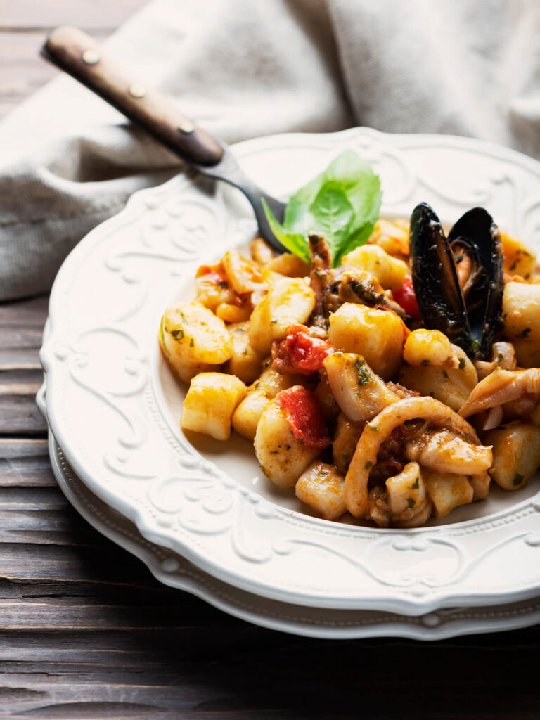 Schräge Seitenansicht eines weißen Tellers mit Reliefrand auf einem zweiten weißen Teller: ein Haufen heller, glatt geschnittener Kartoffel-Gnocchi ohne Rillen mit Tintenfischringen, rechts ragt eine geöffnete Miesmuschel mit beiden schwarzen Schalenhälften heraus, links davon unter dem Basilikum ein dunkler Fangarm und ausgelöstes Muschelfleisch, dazwischen kräftig rote, formstabile Stücke geschälter Tomate und feine Petersilie in dünner, glänzender Sauce, oben ein frischer Basilikumzweig und eine Gabel mit dunklem Holzgriff, dahinter beiges Leinentuch auf dunklem Holz