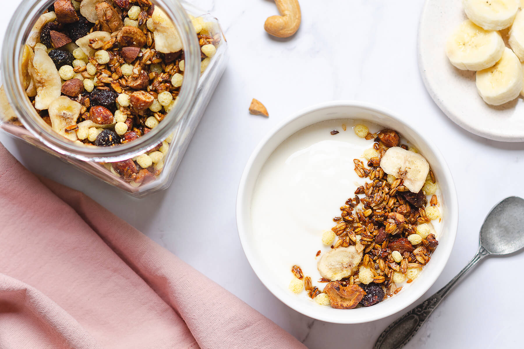 Selbst gemachtes Granola über Naturjoghurt in einer weißen Schale, daneben frische Bananenscheiben und das gefüllte Vorratsglas