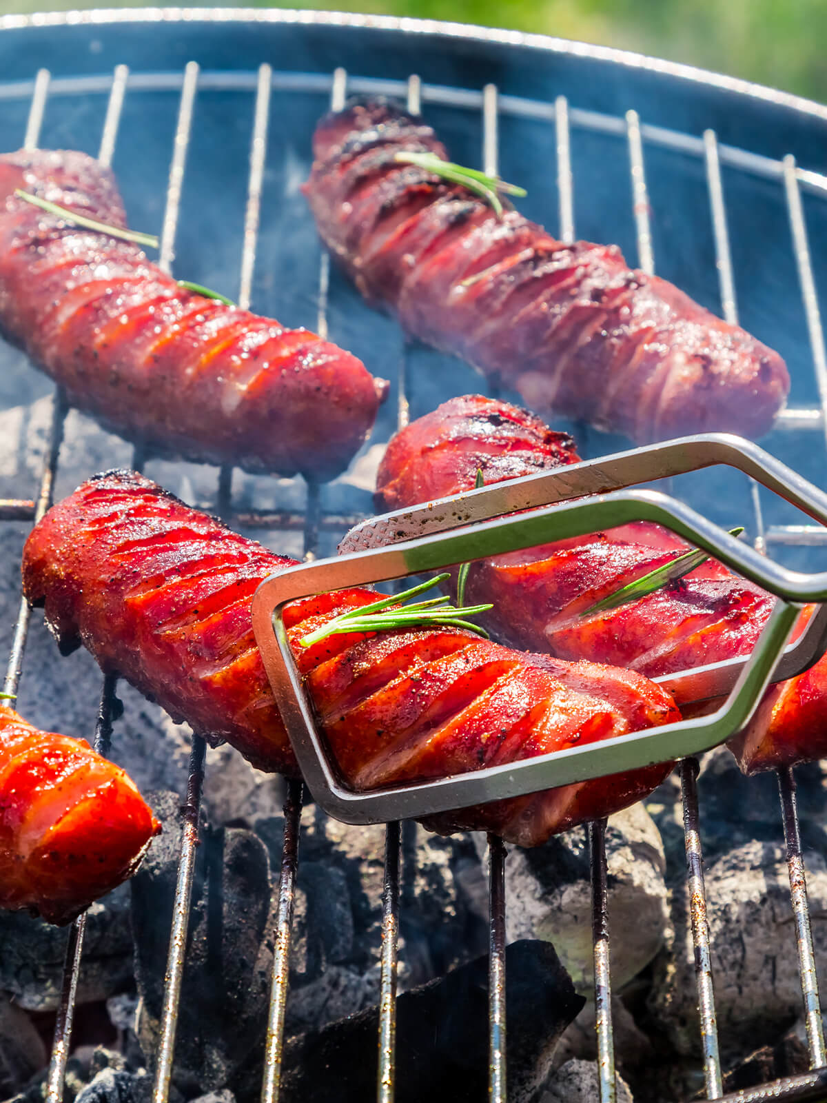 Gegrillte Grillwürste mit roter Paprika-Marinade und Rosmarin auf dem Holzkohlegrill mit Grillzange
