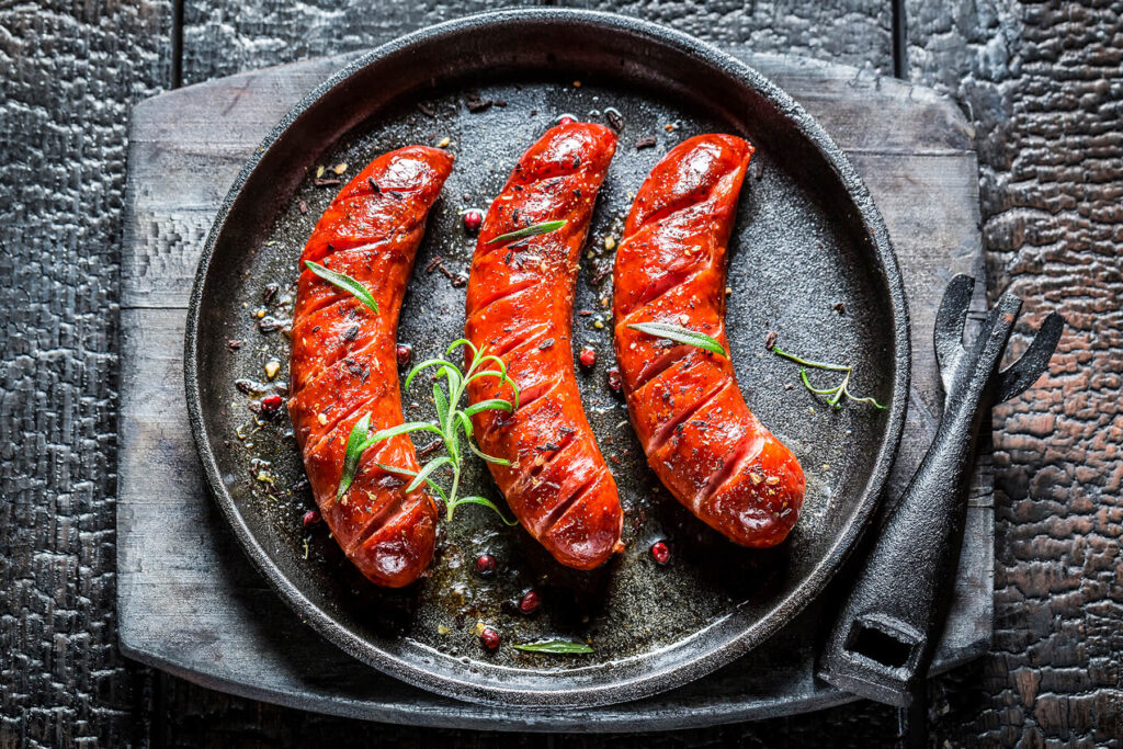 Drei gegrillte Grillwürste mit roter Paprika-Marinade und Rosmarin in einer gusseisernen Pfanne