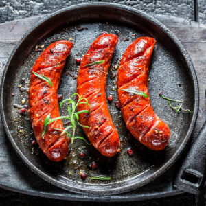 Drei gegrillte Grillwürste mit roter Paprika-Marinade und Rosmarin in einer gusseisernen Pfanne