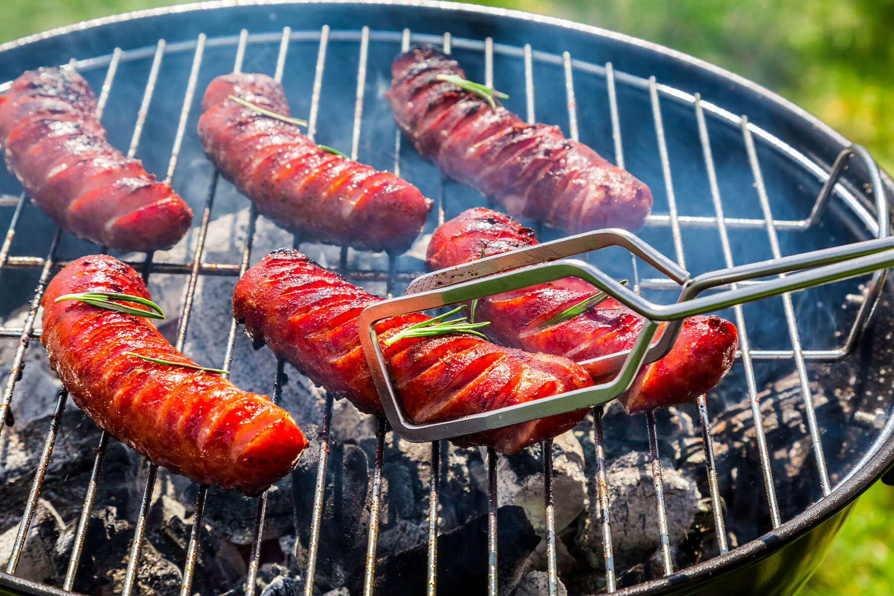 "Rote Grillwürste mit Rosmarin auf dem Holzkohlegrill, die mit einer Grillzange gewendet werden"