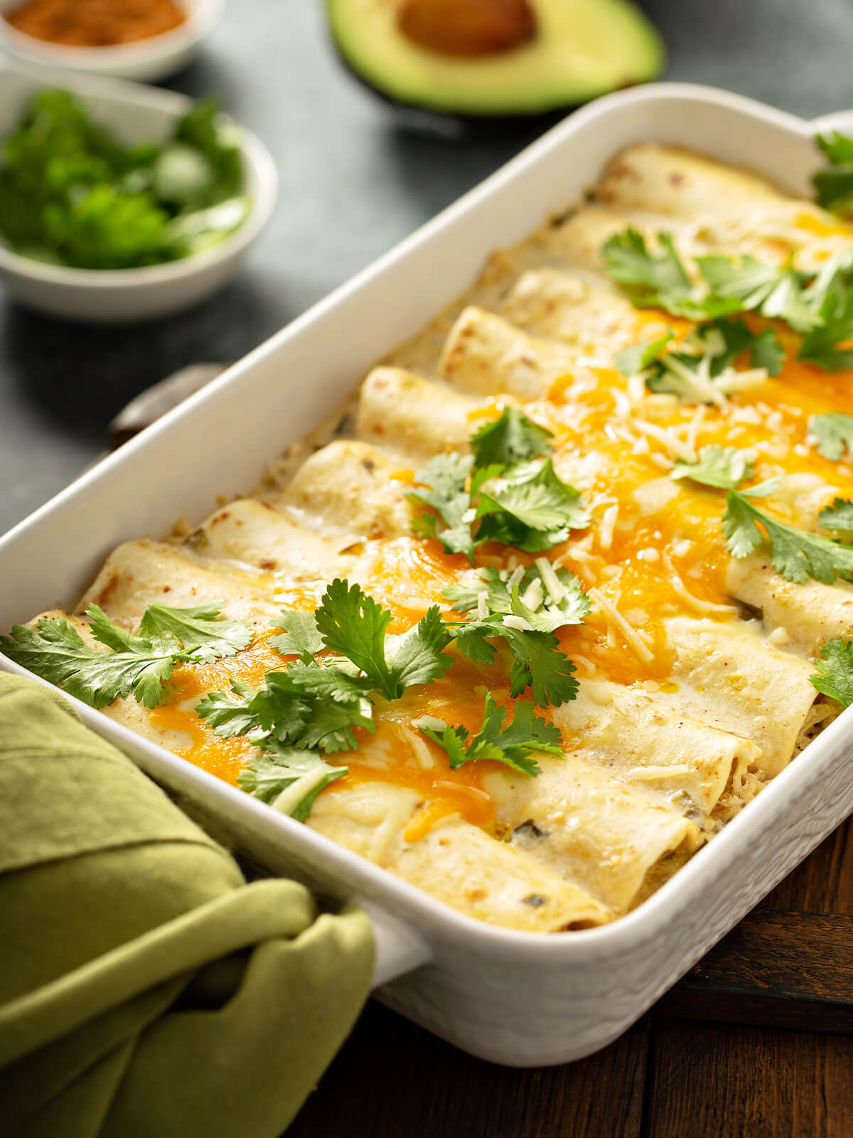 Cremige Hähnchen-Enchiladas in einer weißen Auflaufform, mit weißer Sauce, geschmolzenem Cheddar und frischem Koriander, daneben eine halbe Avocado