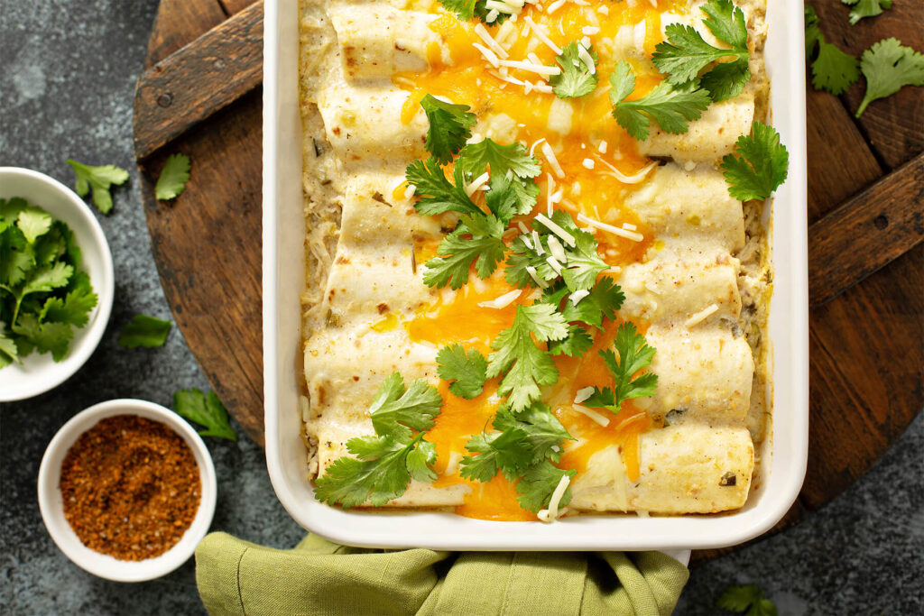 Cremige Hähnchen-Enchiladas von oben in einer weißen Auflaufform, weiße Sauce mit grünen Chilistückchen, geschmolzener Cheddar und frischer Koriander