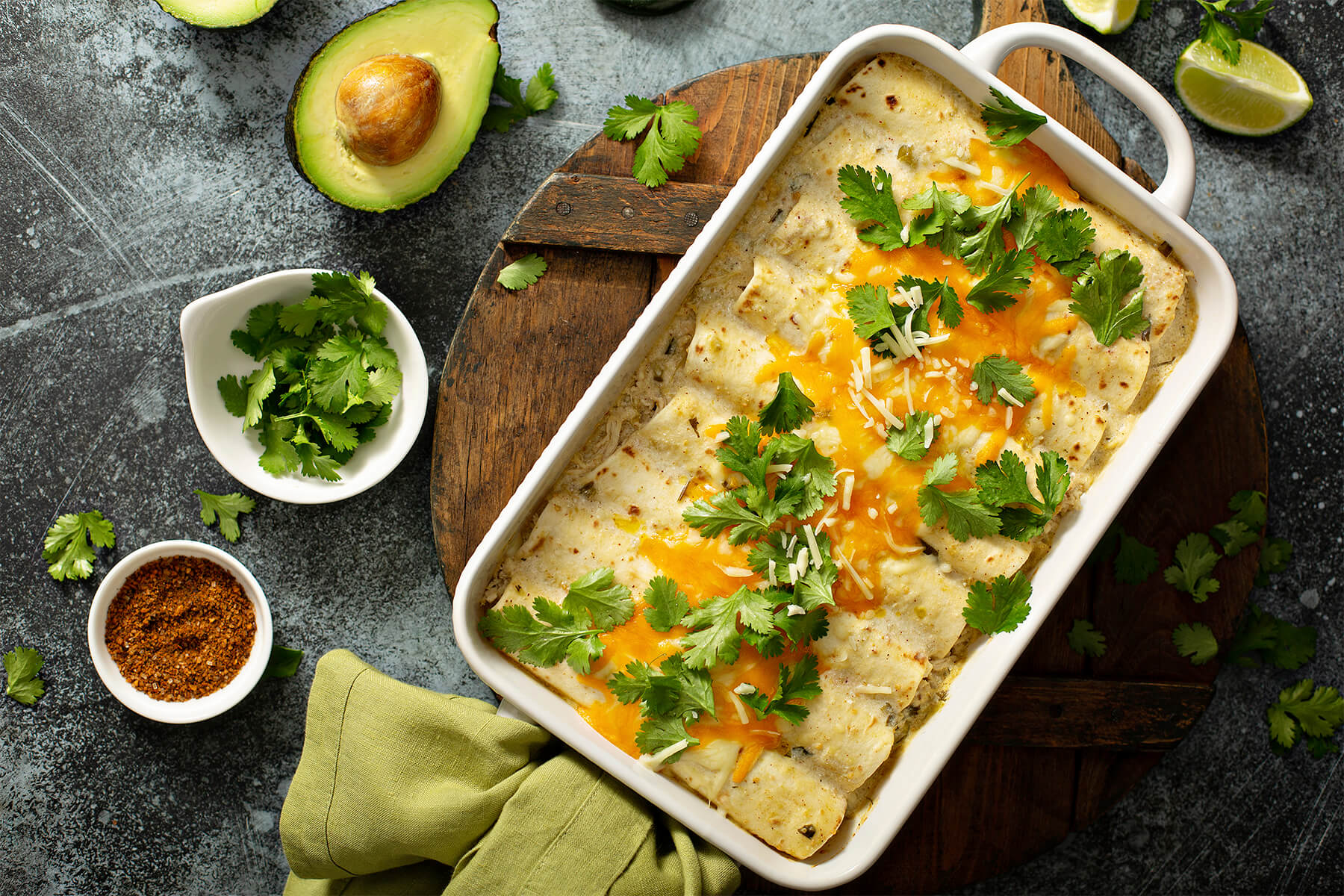 Cremige Hähnchen-Enchiladas von oben in weißer Auflaufform, weiße Sauce mit grünen Chilistückchen, geschmolzener Cheddar und Koriander, daneben Avocado und Limette
