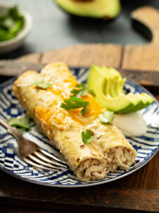 Aufgeschnittene cremige Hähnchen-Enchilada auf einem blau-weißen Teller, Füllung aus zerzupftem Hähnchen in weißer Sauce, dazu Avocado und saure Sahne
