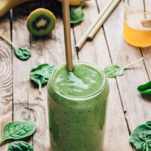 Cremiger grüner Smoothie mit Banane, Kiwi und Spinat im Glas mit Bambus-Trinkhalm, umgeben von Banane und Spinatblättern