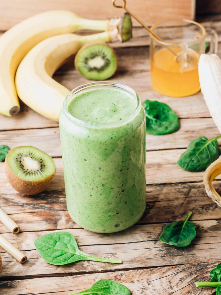 Fertiger grüner Smoothie mit Banane, Kiwi und Spinat im Glas auf einem Holztisch, daneben Banane, Kiwihälften und Spinatblätter