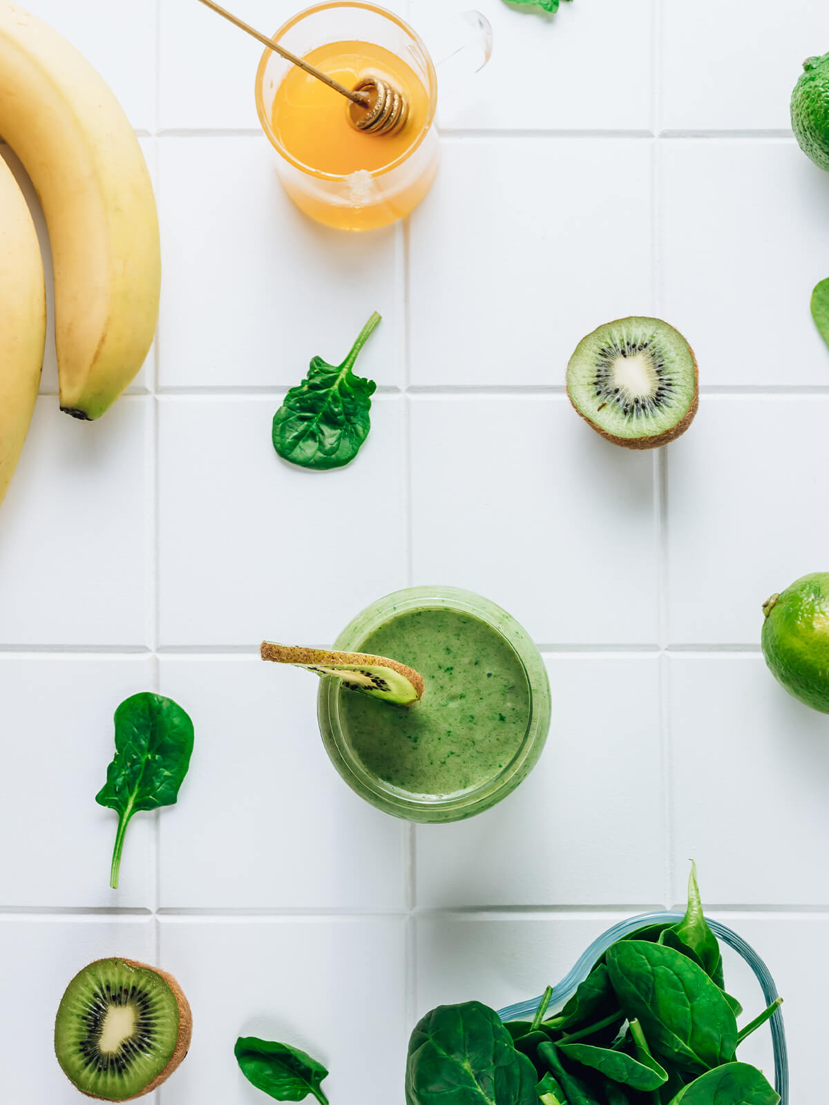 Grüner Smoothie mit Banane, Kiwi und Spinat von oben, umgeben von Bananen, Kiwihälften, Limette und frischen Spinatblättern
