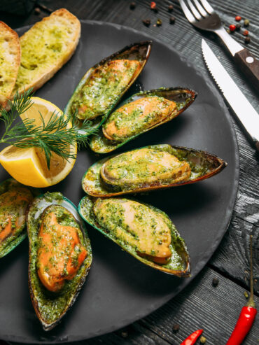 Überbackene Grünschalmuscheln auf der halben Schale mit grüner Kräuter-Knoblauch-Butter auf schwarzem Teller, dazu Knoblauchbrot, Zitrone und Dill