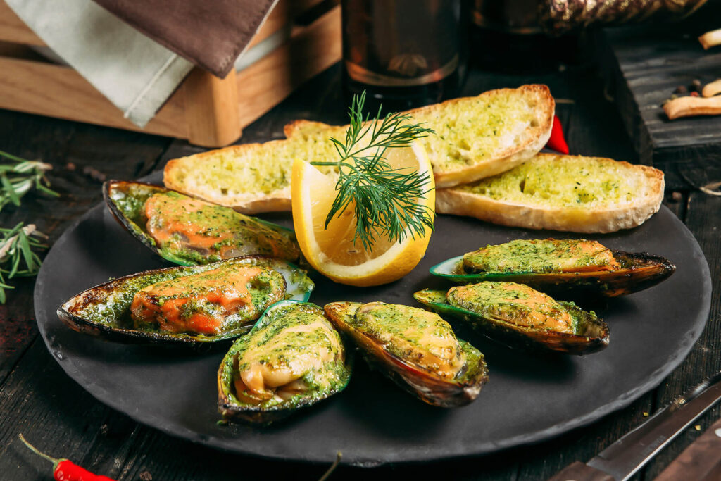 Überbackene Grünschalmuscheln mit grüner Kräuter-Knoblauch-Butter auf einem schwarzen Teller, dazu Knoblauchbrot, Zitrone und Dill