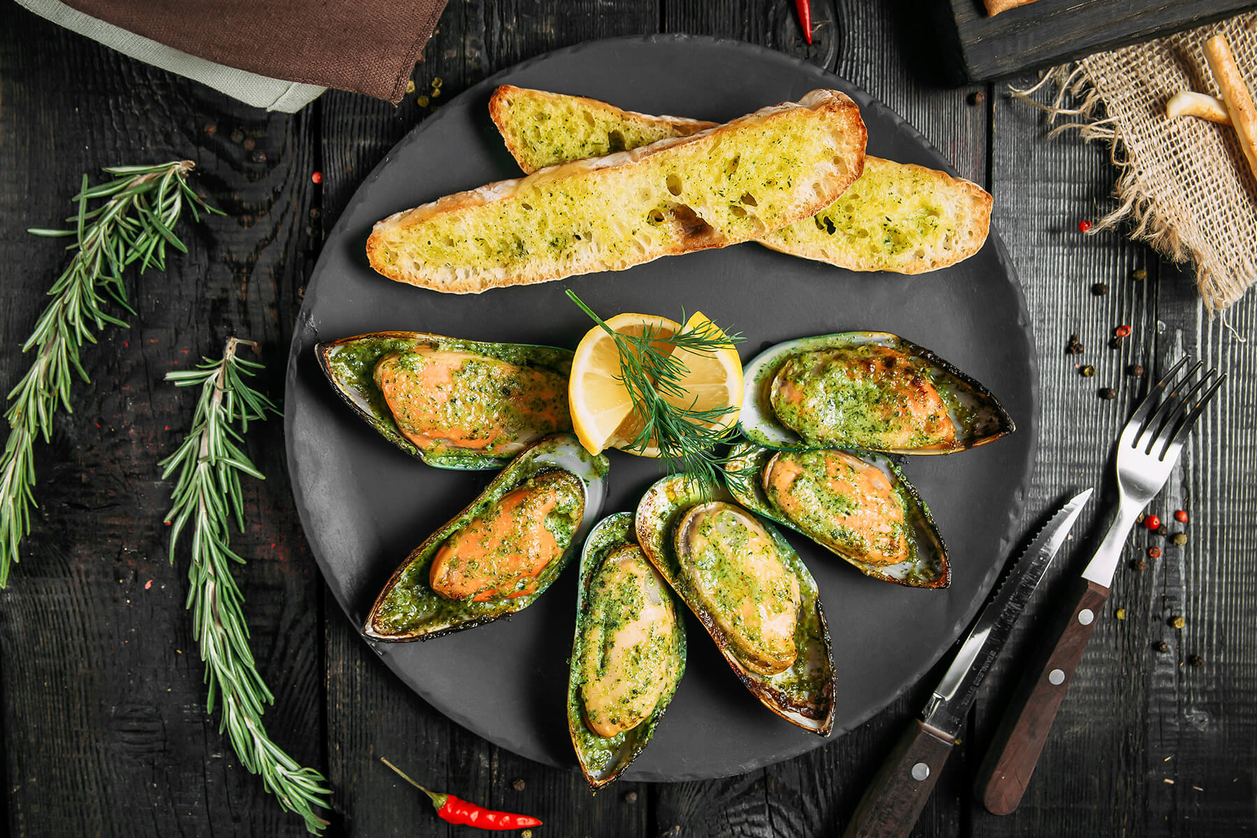 Grünschalmuscheln auf der halben Schale mit grüner Kräuterbutter, angerichtet mit Knoblauchbrot und Zitrone auf schwarzem Teller