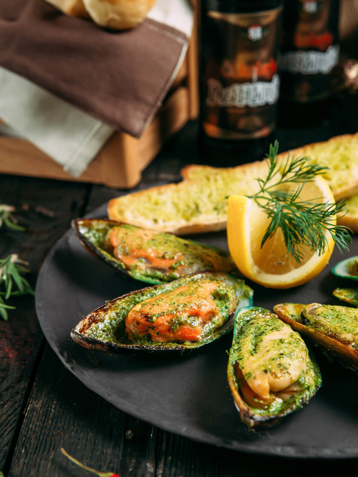 Nahaufnahme überbackener Grünschalmuscheln mit satt grüner Kräuter-Knoblauch-Butter und Knoblauchbrot im Hintergrund