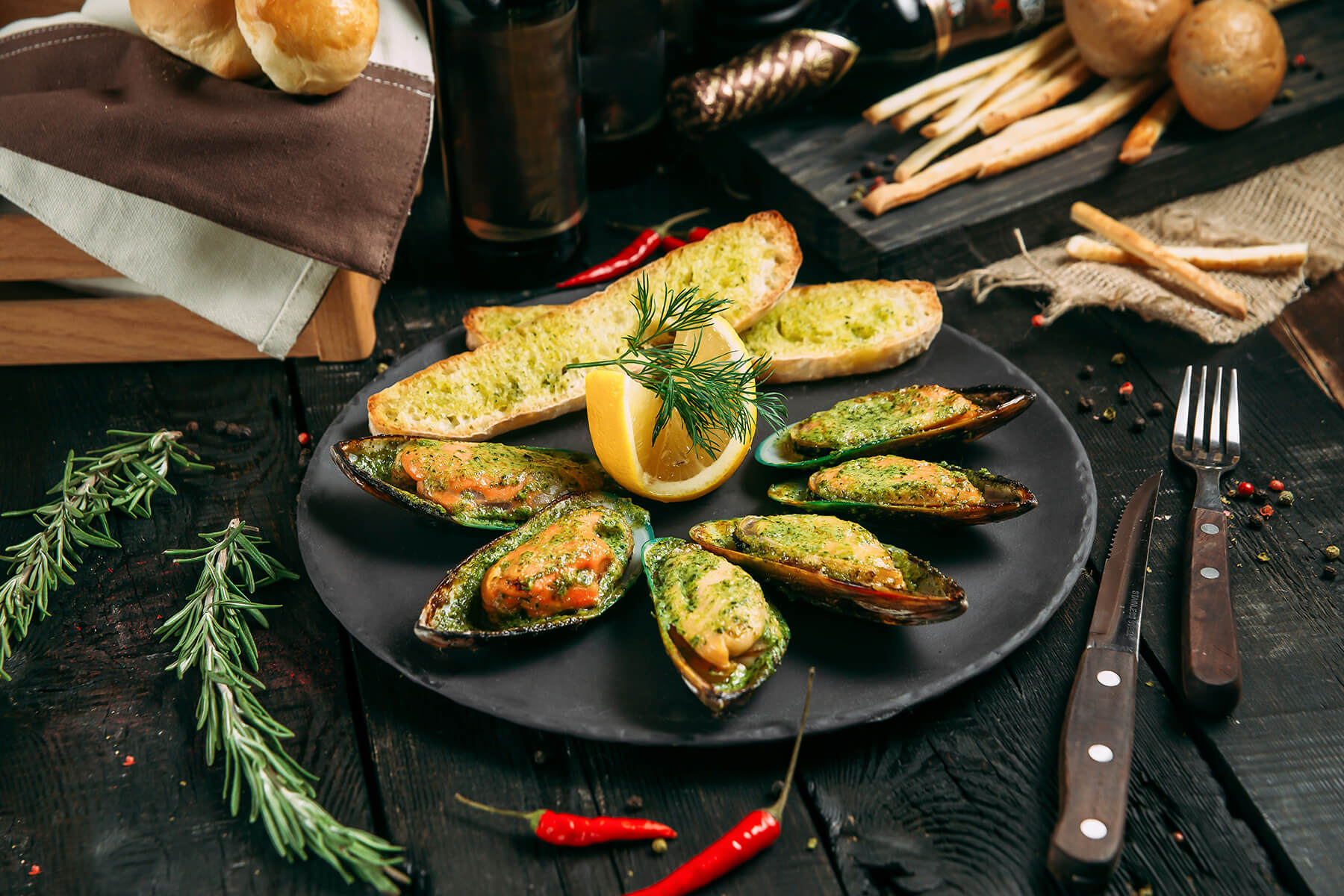 Angerichtete überbackene Grünschalmuscheln mit Knoblauchbrot, Zitrone und Dill auf schwarzem Teller, dazu Rosmarin
