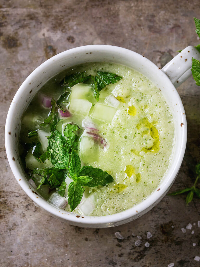 Kalte Gurken-Gazpacho mit Minze in einer weißen Tasse, garniert mit Gurkenwürfeln, roten Zwiebeln, frischer Minze und Olivenöl