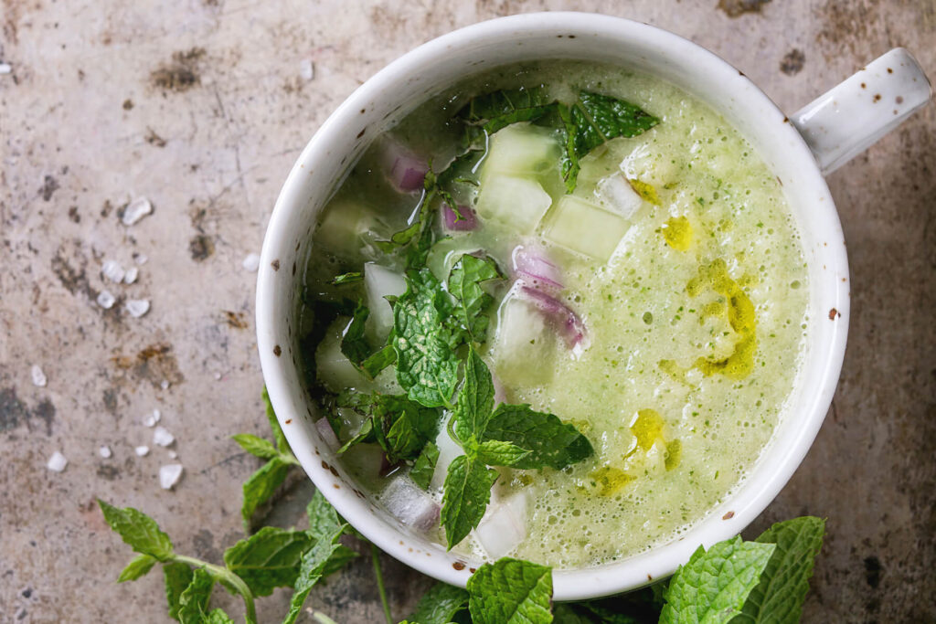 Erfrischende kalte Gurken-Gazpacho mit Minze in einer weißen Tasse, mit Gurken, roter Zwiebel und Olivenöl garniert, daneben frische Minze