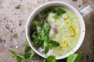 Erfrischende kalte Gurken-Gazpacho mit Minze in einer weißen Tasse, mit Gurken, roter Zwiebel und Olivenöl garniert, daneben frische Minze