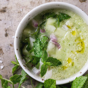 Erfrischende kalte Gurken-Gazpacho mit Minze in einer weißen Tasse, mit Gurken, roter Zwiebel und Olivenöl garniert, daneben frische Minze