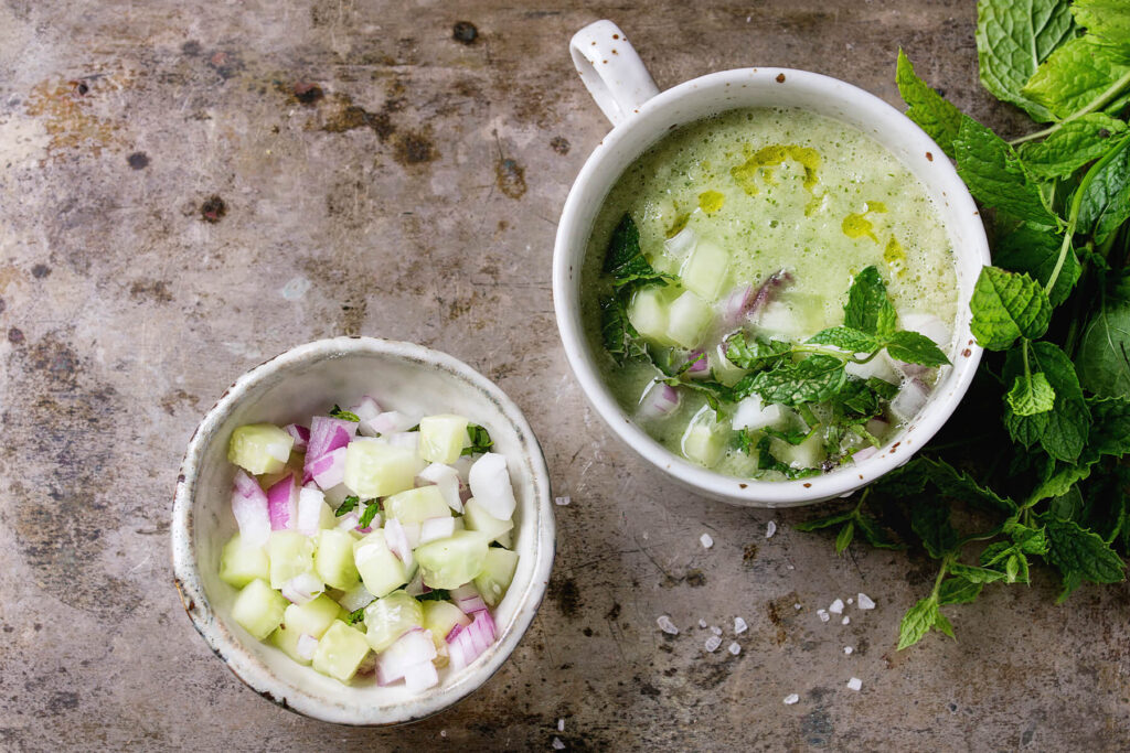 Kalte Gurken-Gazpacho von oben in einer Tasse, daneben eine kleine Schale mit gewürfelter Gurke und roter Zwiebel sowie frische Minze