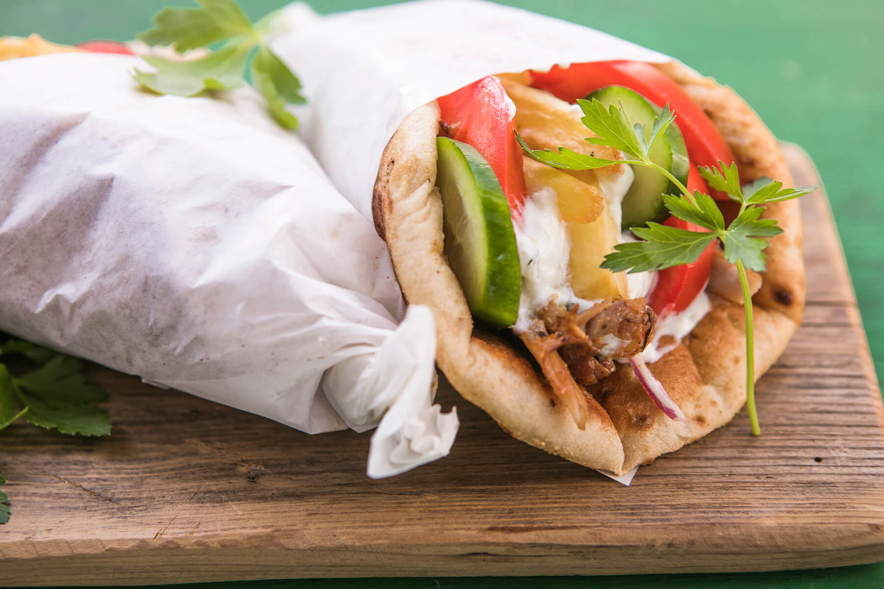 Ein in weißes Papier gewickeltes griechisches Pita-Brot auf einem rustikalen Holzbrett vor grünem Untergrund; aus der offenen Seite schauen rote Tomatenspalten, grüne Gurkenscheiben, dicke hellgoldene Pommes frites, weiße Joghurtsauce, dunkel gebratene Fleischstreifen, rote Zwiebeln und glatte Petersilie heraus