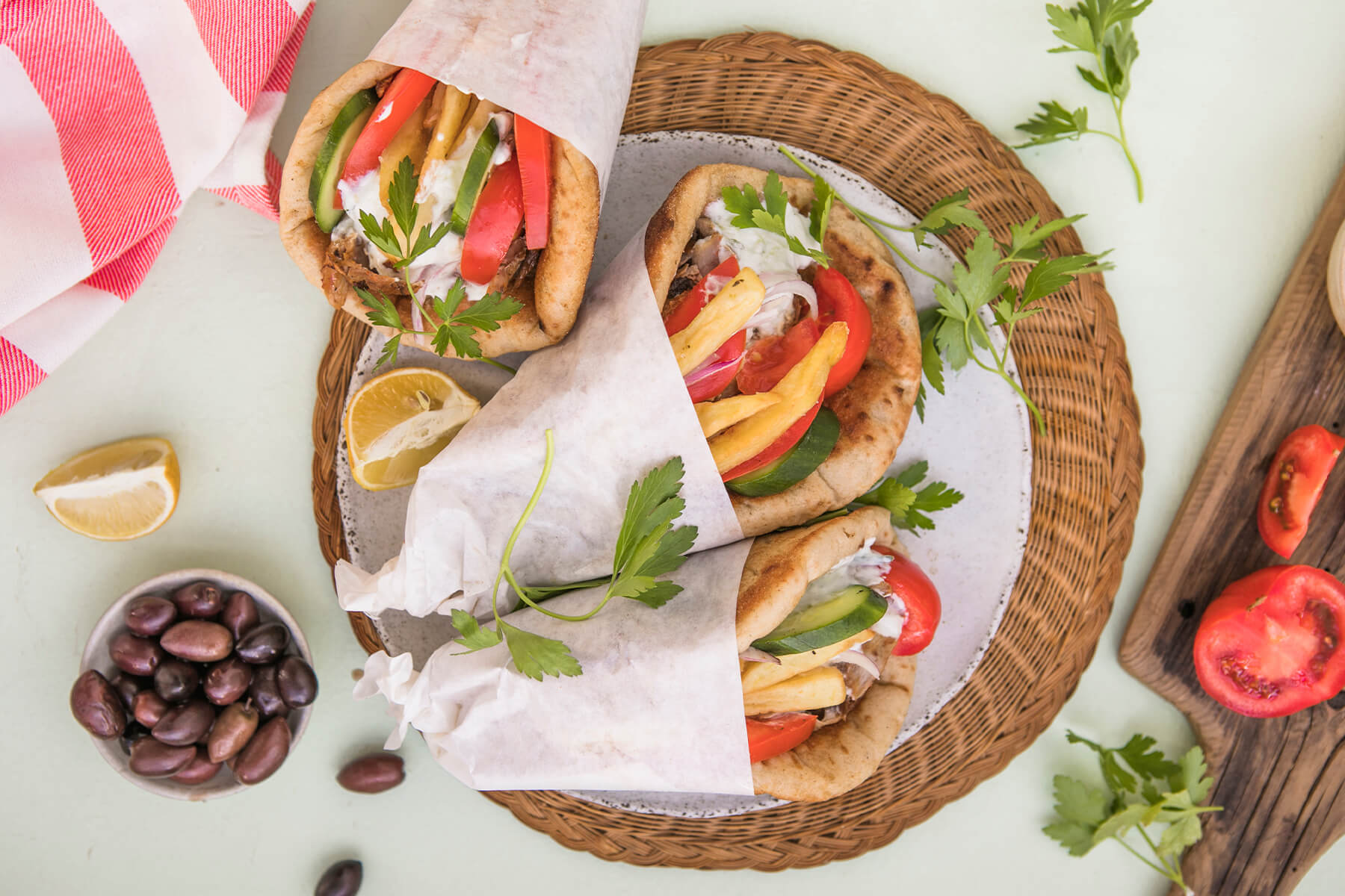 Drei in weißes Papier gewickelte griechische Pita-Brote von oben auf einem hellen, gesprenkelten Teller auf runder Korbmatte; sichtbar sind rote Tomatenspalten, Gurkenscheiben, hellgoldene Pommes frites, rote Zwiebelringe, weiße Joghurtsauce und glatte Petersilie; daneben ein Schälchen mit dunklen Kalamata-Oliven, Zitronenspalten, ein Holzbrett mit halbierten Tomaten und ein rot-weiß gestreiftes Tuch