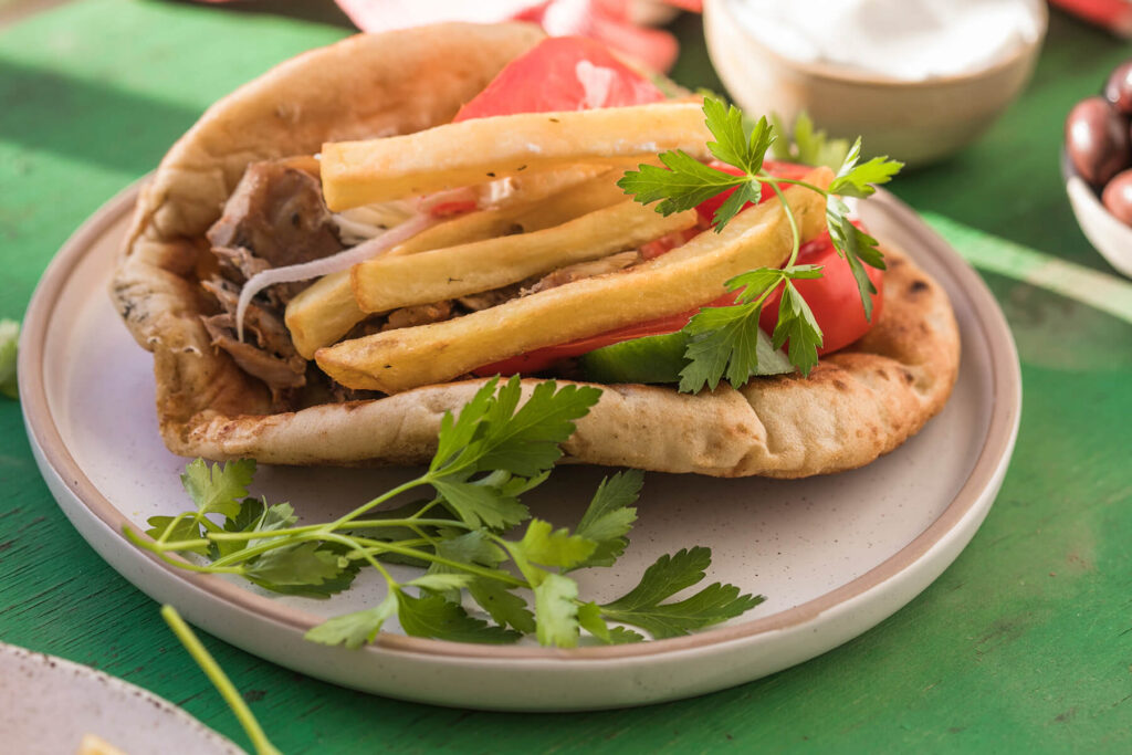 Nahaufnahme eines gefüllten griechischen Pita-Brots auf einem hellen, gesprenkelten Teller mit rosé Rand: dunkel gebratene Fleischstreifen, rote Zwiebelringe, dicke hellgoldene Pommes frites, rote Tomatenspalten, eine Gurkenscheibe und glatte Petersilie; grüner Holztisch, im Hintergrund unscharf ein Schälchen Joghurt und dunkle Oliven