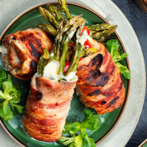Drei Hähnchen-Spargel-Röllchen im Baconmantel auf grünem Teller, grüne Spargelspitzen mit Frischkäse schauen heraus, auf Feldsalat angerichtet