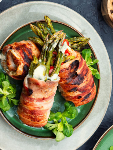 Drei Hähnchen-Spargel-Röllchen im Baconmantel auf grünem Teller, grüne Spargelspitzen mit Frischkäse schauen heraus, auf Feldsalat angerichtet