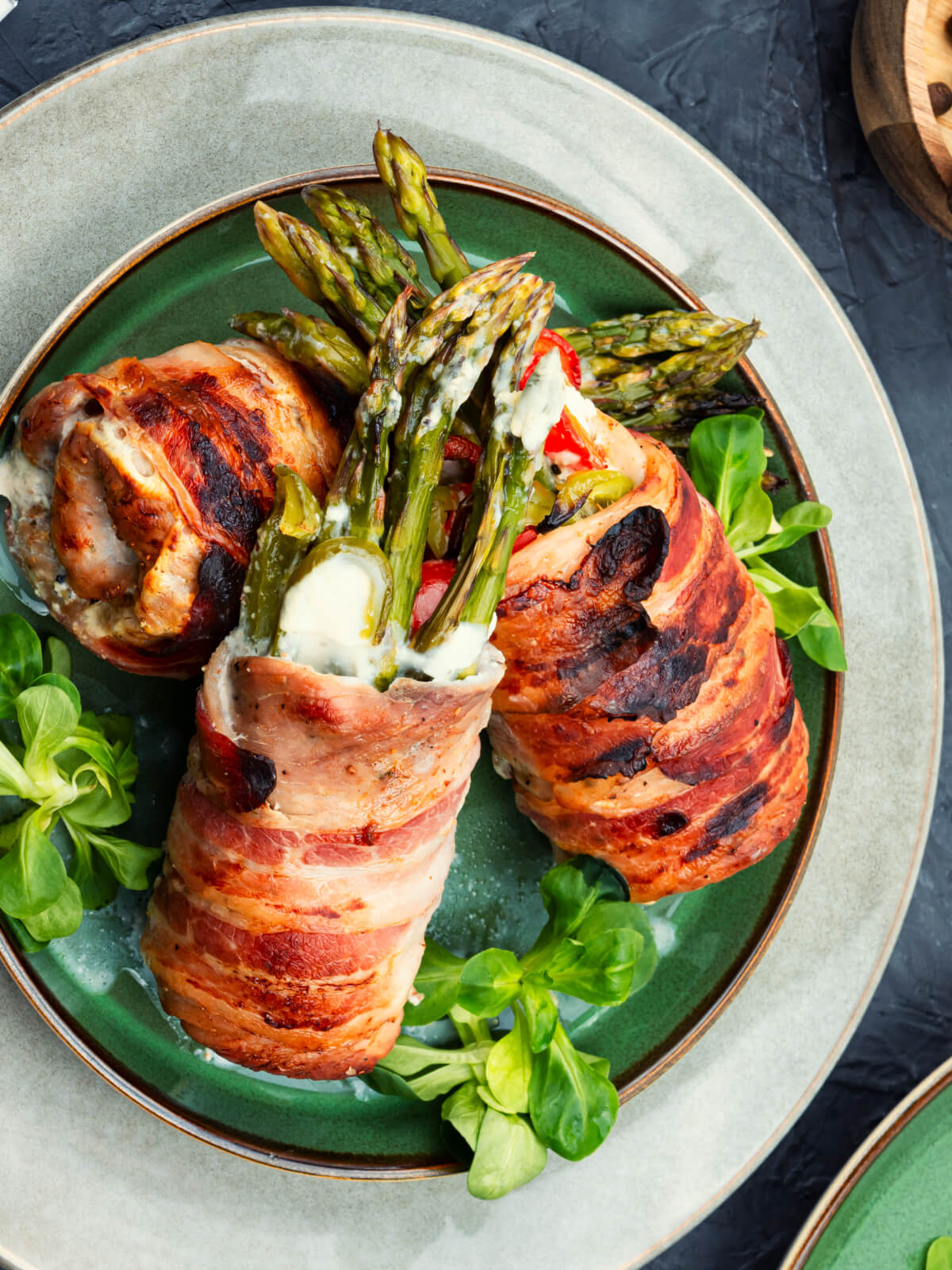 Drei Hähnchen-Spargel-Röllchen im Baconmantel auf grünem Teller, grüne Spargelspitzen mit Frischkäse schauen heraus, auf Feldsalat angerichtet