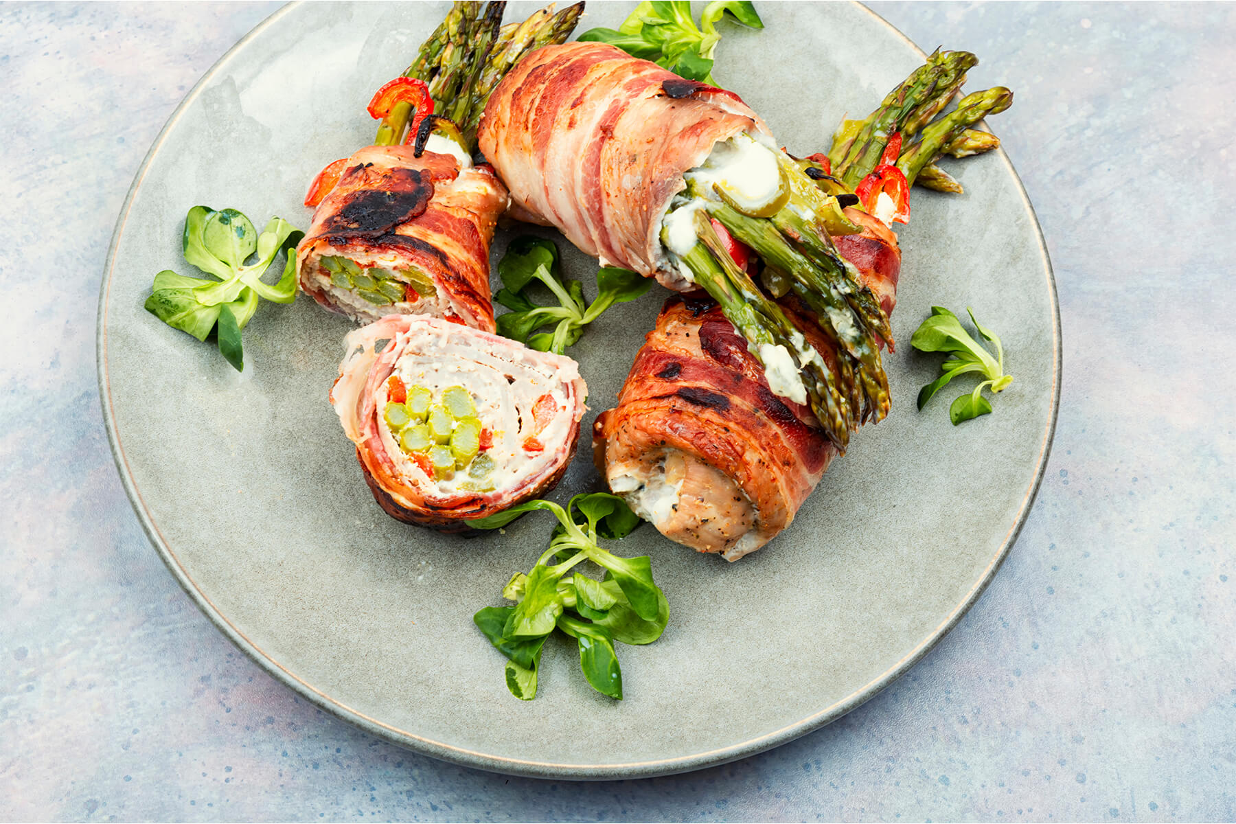 Hähnchen-Spargel-Röllchen im Baconmantel auf einem Teller, ein Röllchen aufgeschnitten zeigt die Spirale aus Hähnchen, Frischkäse, Spargel und Paprika