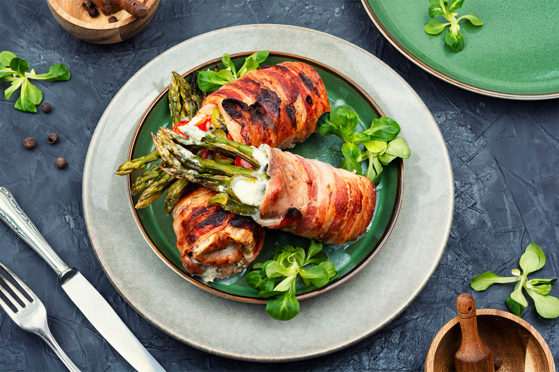 Zwei Hähnchen-Spargel-Röllchen im Baconmantel auf grünem Teller mit Feldsalat und Besteck auf dunklem Hintergrund