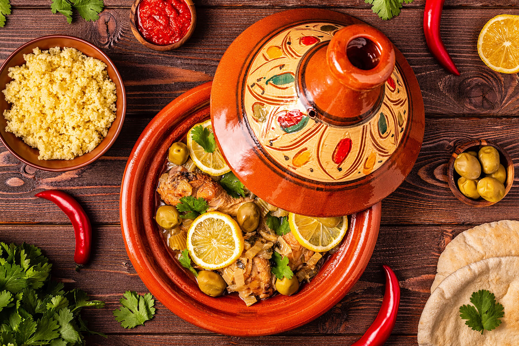 Flatlay mit marokkanischer Hähnchen-Tajine, einer Schale Couscous, Harissa, grünen Oliven, Fladenbrot und frischer Chili