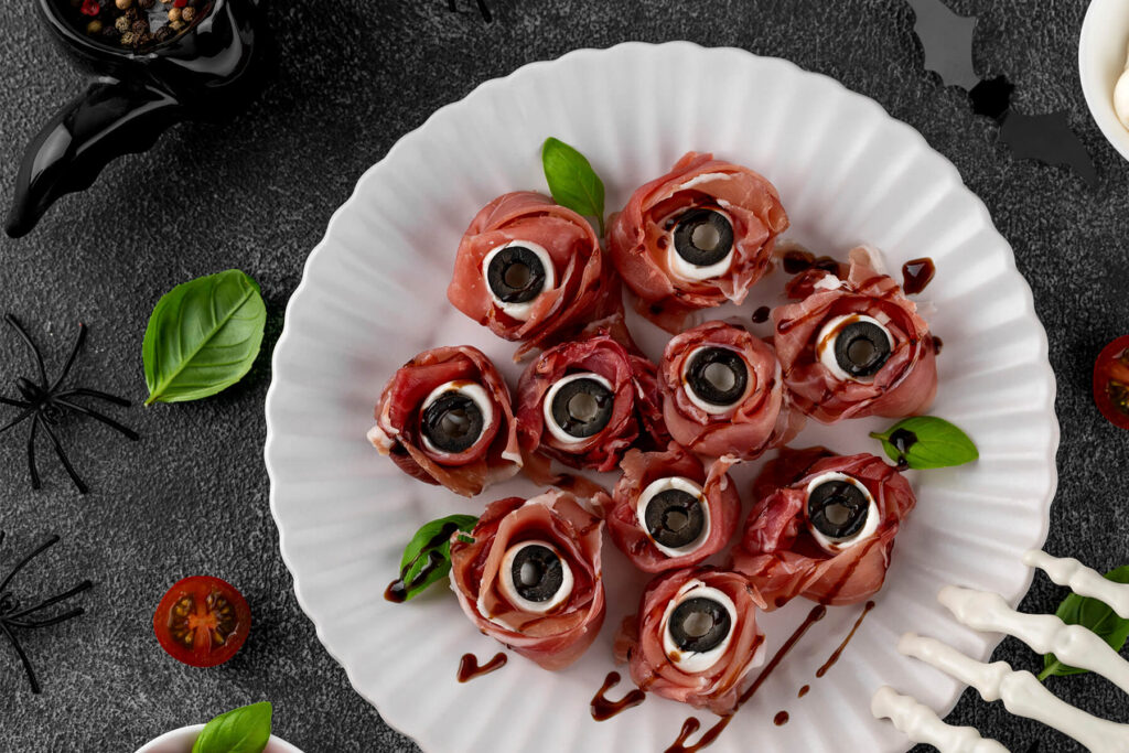 Mehrere Halloween-Augäpfel aus rosa Prosciutto-Röschen mit weißer Mozzarella-Kugel und schwarzer Olivenscheibe als Pupille auf einem weißen geriffelten Teller, überzogen mit feinen dunklen Balsamico-Äderchen und mit Basilikum garniert; dunkler Schiefergrund mit schwarzer Deko-Spinne, Fledermaus und einer weißen Skeletthand am Rand