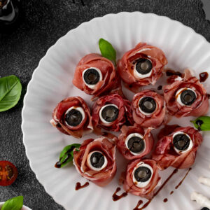 Mehrere Halloween-Augäpfel aus rosa Prosciutto-Röschen mit weißer Mozzarella-Kugel und schwarzer Olivenscheibe als Pupille auf einem weißen geriffelten Teller, überzogen mit feinen dunklen Balsamico-Äderchen und mit Basilikum garniert; dunkler Schiefergrund mit schwarzer Deko-Spinne, Fledermaus und einer weißen Skeletthand am Rand