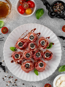 Fertige Halloween-Augäpfel aus Prosciutto-Röschen mit weißer Mozzarella-Kugel und schwarzer Olivenscheibe als Pupille, mit dunklen Balsamico-Äderchen beträufelt und mit Basilikum garniert, auf einem weißen geriffelten Teller; heller Steingrund mit Kirschtomaten, Pfefferkörnern und Halloween-Deko