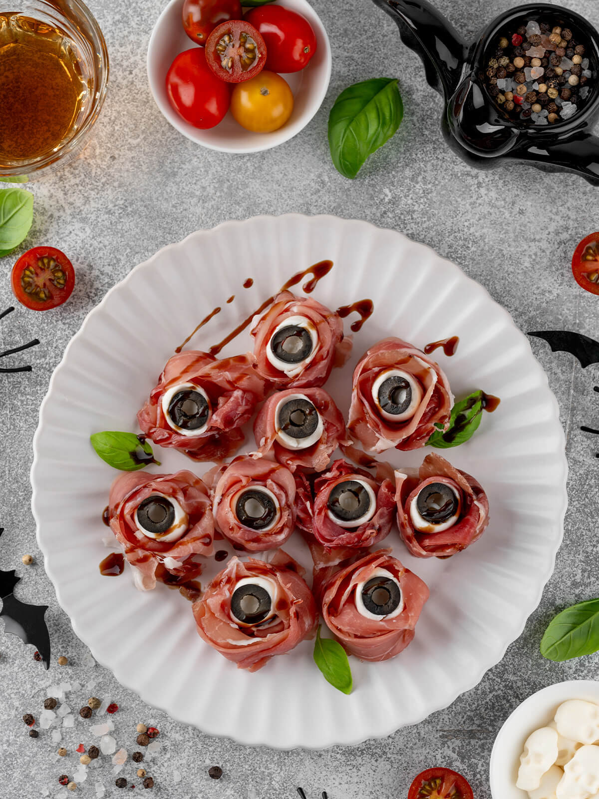 Fertige Halloween-Augäpfel aus Prosciutto-Röschen mit weißer Mozzarella-Kugel und schwarzer Olivenscheibe als Pupille, mit dunklen Balsamico-Äderchen beträufelt und mit Basilikum garniert, auf einem weißen geriffelten Teller; heller Steingrund mit Kirschtomaten, Pfefferkörnern und Halloween-Deko