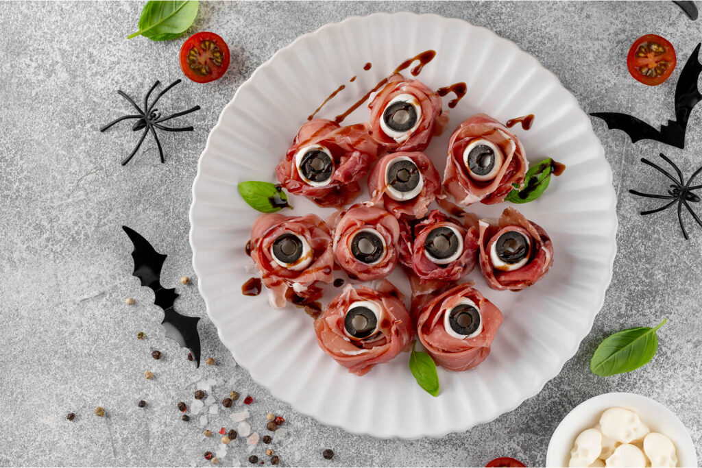 Mehrere Halloween-Augäpfel aus rosa Prosciutto-Röschen mit weißer Mozzarella-Kugel und schwarzer Olivenscheibe als Pupille, mit dunklen Balsamico-Linien überzogen und mit Basilikum garniert, auf einem weißen geriffelten Teller; heller Grund mit schwarzen Deko-Spinnen, Fledermäusen und einer kleinen Schale Mozzarella-Totenköpfe