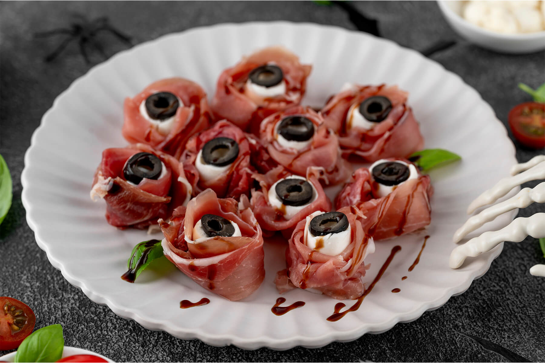 Nahaufnahme mehrerer Halloween-Augäpfel aus rosa Prosciutto-Röschen mit weißer Mozzarella-Kugel und schwarzer Olivenscheibe als Pupille, überzogen mit dunklen Balsamico-Äderchen, auf einem weißen geriffelten Teller; dunkler Grund mit Basilikum, einer halbierten Kirschtomate und einer weißen Skeletthand am Tellerrand