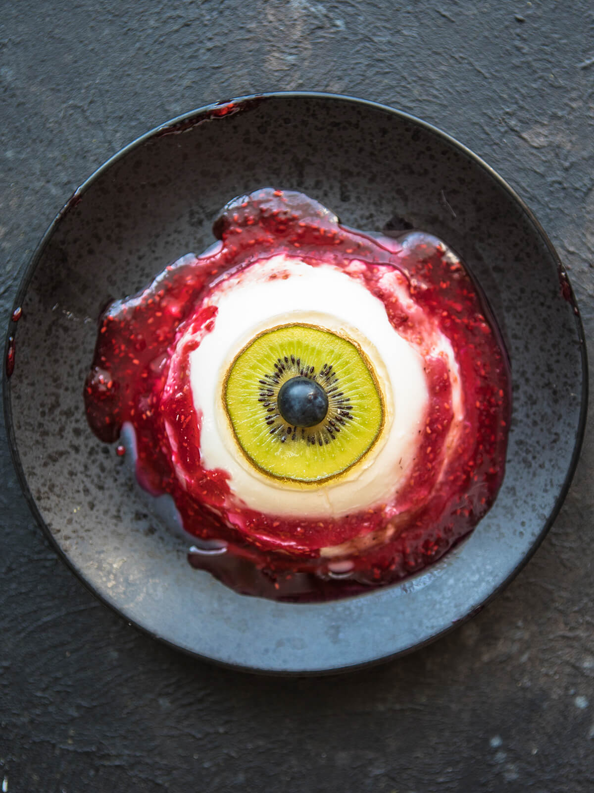 Blutige Augapfel-Panna-Cotta von oben mit Kiwi-Iris, Heidelbeer-Pupille und Himbeersauce als Blut