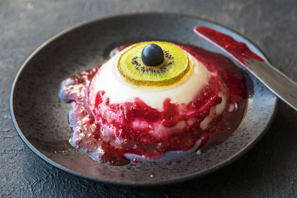 Blutige Augapfel-Panna-Cotta mit Kiwi-Iris und Heidelbeer-Pupille, Himbeersauce läuft über die Schulter der Kuppel