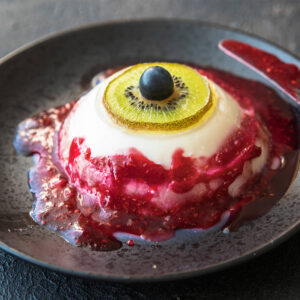 Blutige Augapfel-Panna-Cotta mit Kiwi-Iris und Heidelbeer-Pupille, Himbeersauce läuft über die Schulter der Kuppel