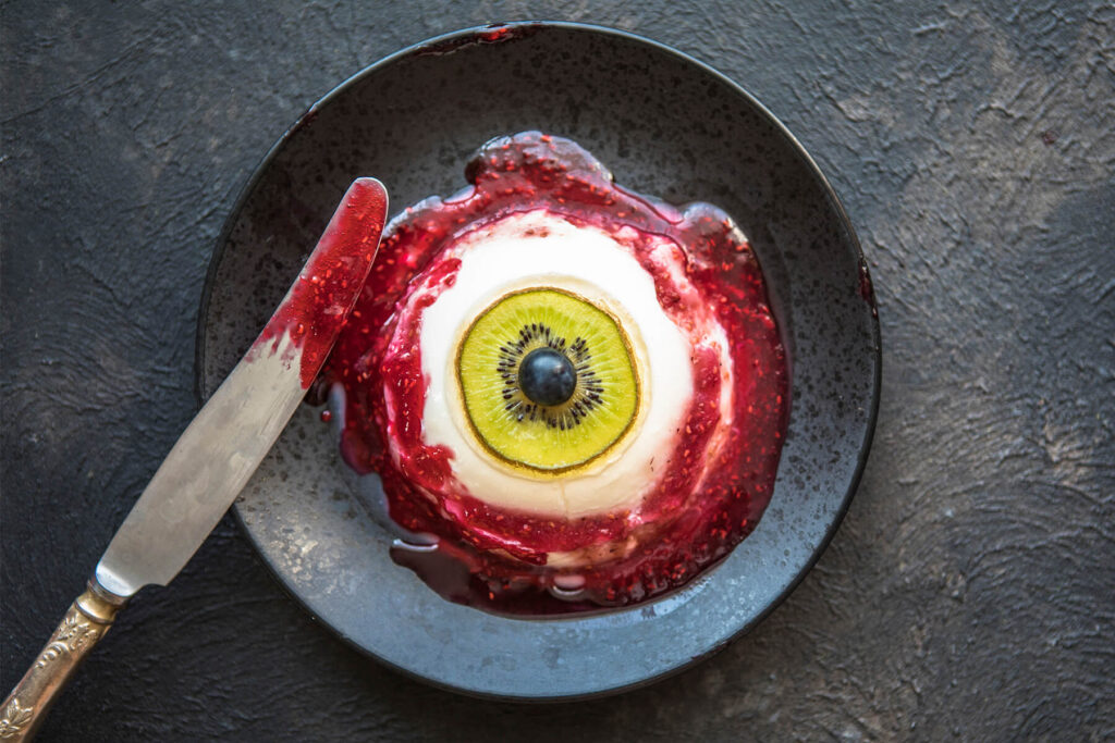 Augapfel-Panna-Cotta von oben auf dunklem Teller, dickes Himbeer-Blut mit Kernen ringsum, daneben ein Messer mit Sauce