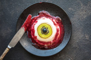 Augapfel-Panna-Cotta von oben auf dunklem Teller, dickes Himbeer-Blut mit Kernen ringsum, daneben ein Messer mit Sauce